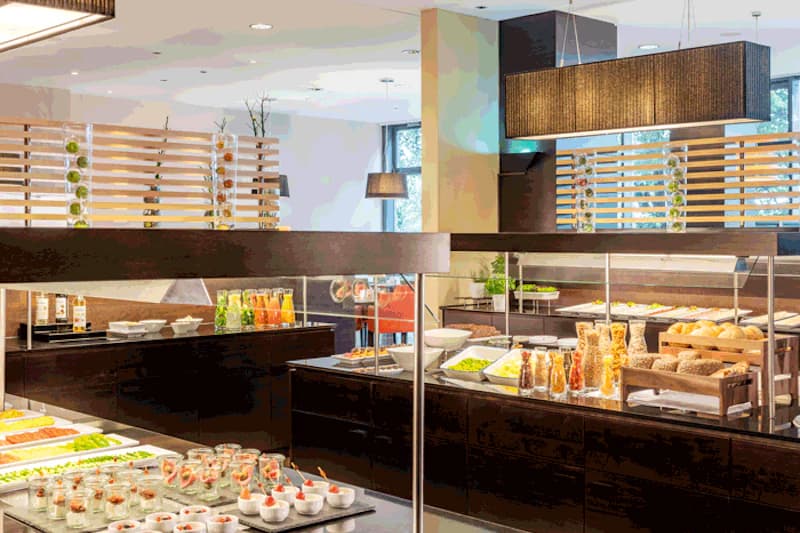 Buffet mit Brötchen, Cerealien, kleinen Dessertgläsern, Säften und Salaten in modernem Frühstücksraum.