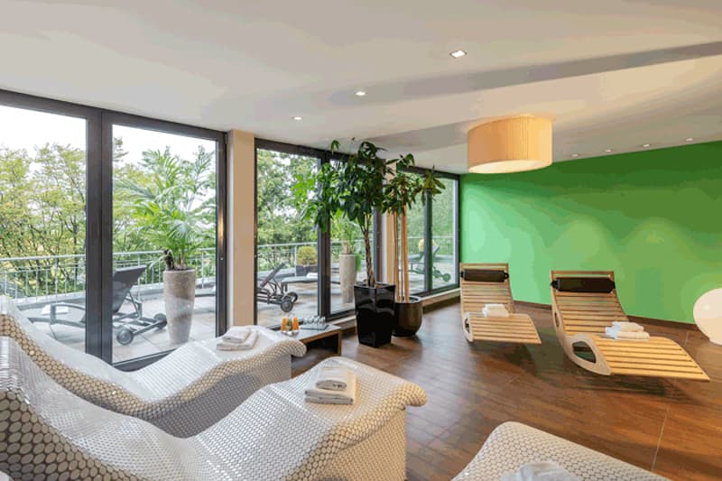 Ruheraum mit geschwungenen Liegen, Holzliegen, grüner Wand und großen Fensterfronten zur Terrasse.
