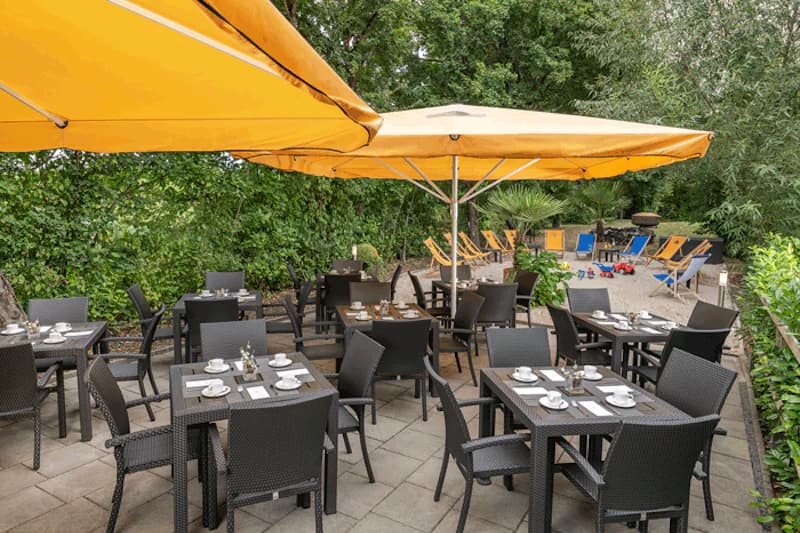 Freiluftterrasse mit gedeckten Tischen, gelben Sonnenschirmen und Liegestühlen vor grünem Bewuchs.