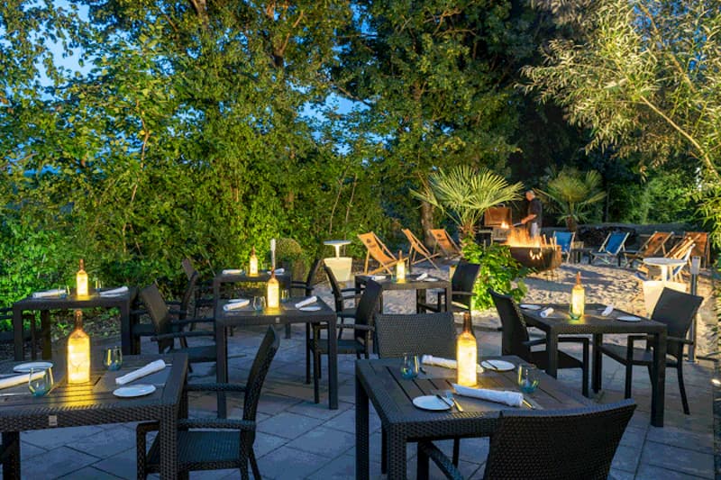 Beleuchtete Hotelterrasse mit gedeckten Tischen, Stühlen, leuchtenden Flaschen, Liegestühlen und Feuerstelle am Baumbestand.