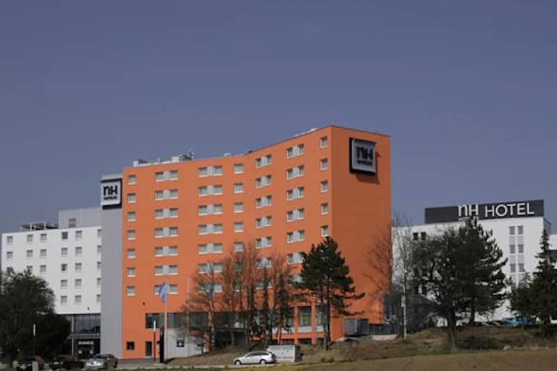 Oranges mehrstöckiges NH Hotel mit weißem Nebengebäude, Bäumen und parkenden Autos im Vordergrund.