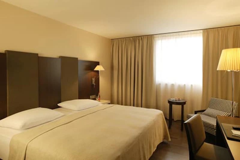Hotelzimmer mit großem Doppelbett, dunklem Holzkopfteil, Nachttischlampen, Fenster mit Vorhängen und Sitzbereich.