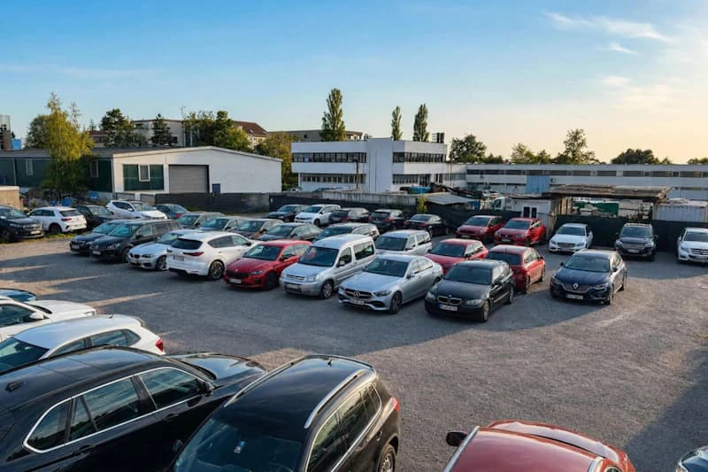 Schotterparkplatz mit zahlreichen geparkten Autos in Reihen, niedrige Hallen und Bäume im Hintergrund.