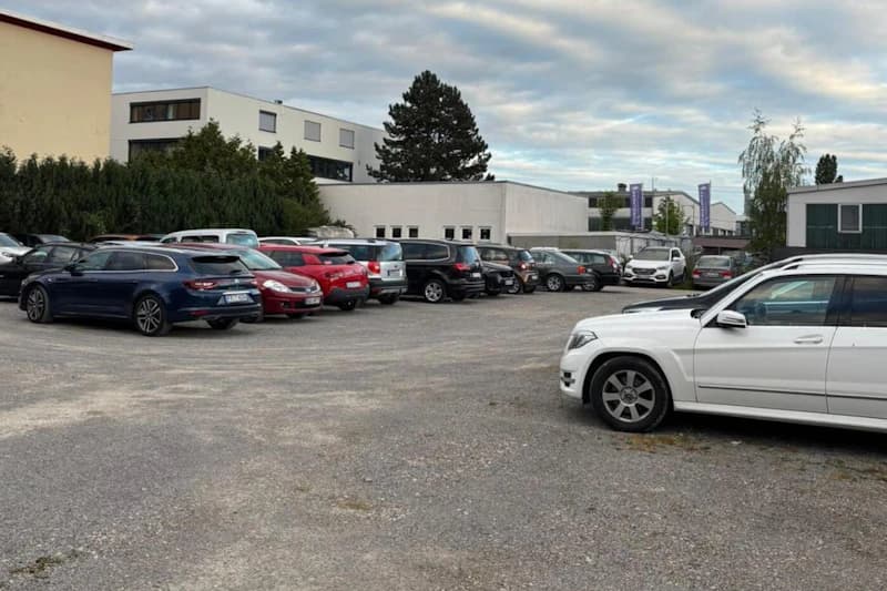 Großer Kiesparkplatz mit mehreren geparkten Autos, niedrige Gewerbebauten und bewölkter Himmel im Hintergrund.