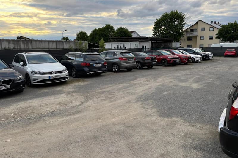 Großer Schotterparkplatz mit einer Reihe geparkter Autos entlang einer Umzäunung, Gebäude und Wohnhäuser im Hintergrund unter wolkigem Himmel.
