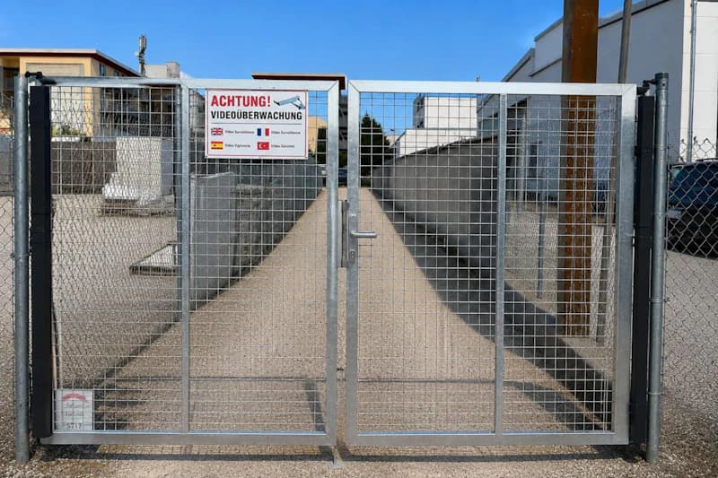 Metallgittertor mit Schloss und Schild Achtung Videoüberwachung vor einem gepflasterten Durchgang.