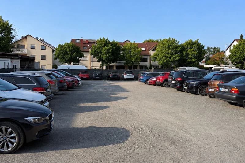 Geparkte Autos auf Schotterparkplatz, Reihen links und rechts, Wohnhäuser und Bäume im Hintergrund.