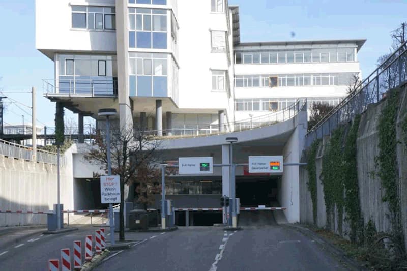 Einfahrt zu Parkhaus mit zwei Schranken, Leuchtsignalen, Bürogebäude darüber und Beleuchtung.