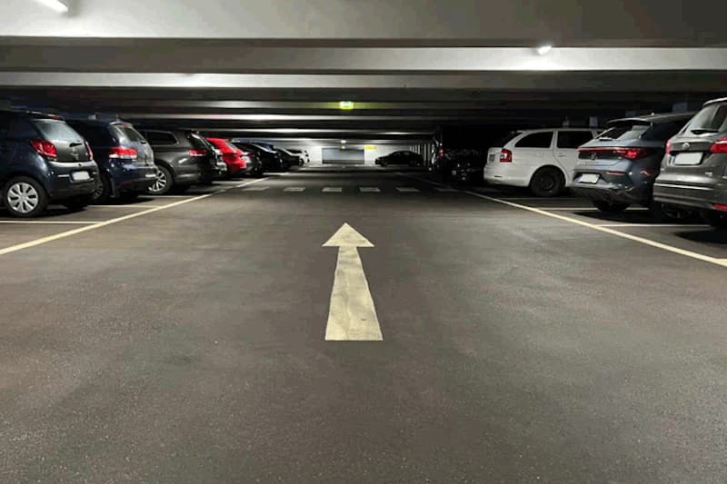 Beleuchtetes Parkhaus mit großem Richtungspfeil auf der Fahrbahn und Autos beidseits geparkt.