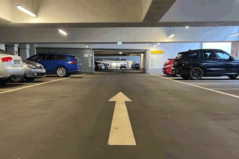 Innenraum eines mehrstöckigen Parkhauses mit weissem Richtungspfeil auf dem Boden und geparkten Autos an den Seiten.