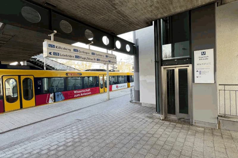 Gelbe Stadtbahn am Bahnsteig, überdachte Haltestelle mit Aufzug und Richtungstafel unter Betonlage.