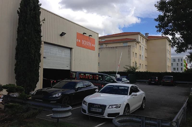 Parkplatz vor Lagerhalle mit orangem BOXX'IN Schild, mehrere geparkte Autos, Wohnhaus im Hintergrund, bewölkter Himmel.