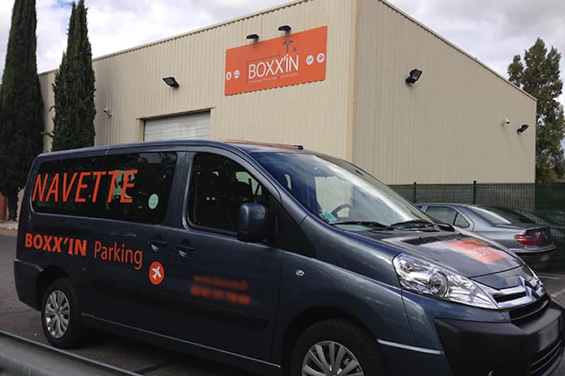 Dunkelgrauer Shuttle vor heller Halle mit orangefarbenem BOXX'IN-Logo an der Fassade.