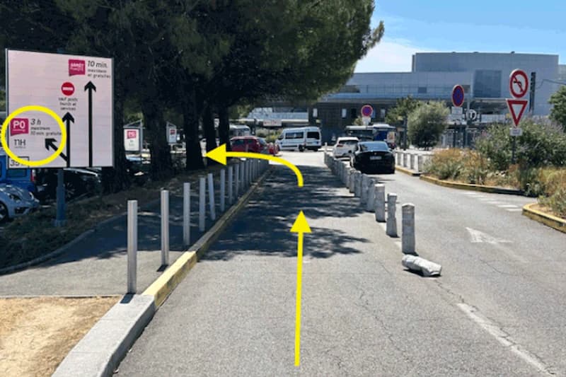 Fahrbahn mit beidseitigen Pollern, Bäumen links, mehreren stehenden Autos und Terminalgebäude im Hintergrund.