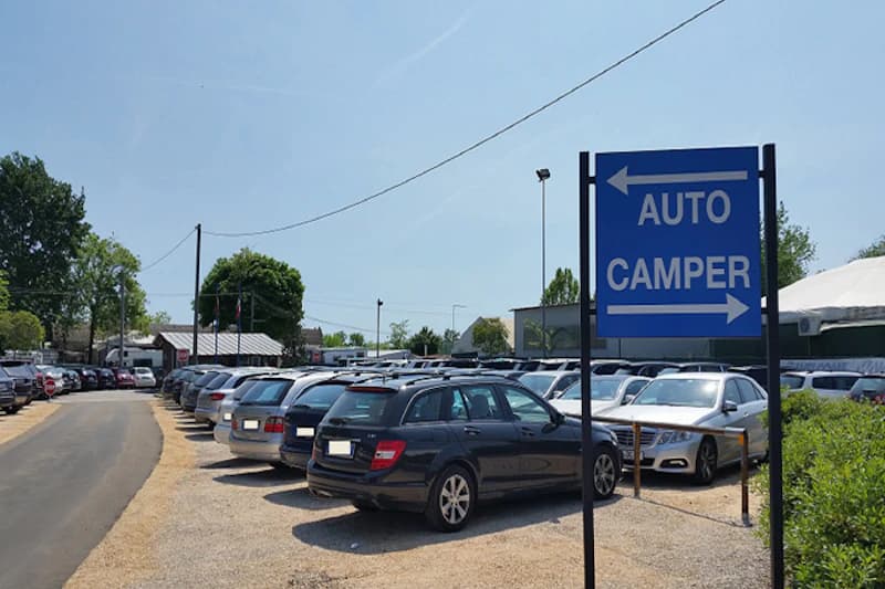 Umzäunter, beleuchteter Parkplatz mit dicht geparkten Autos auf Kies und einem Wegweiser für Pkw und Wohnmobile.