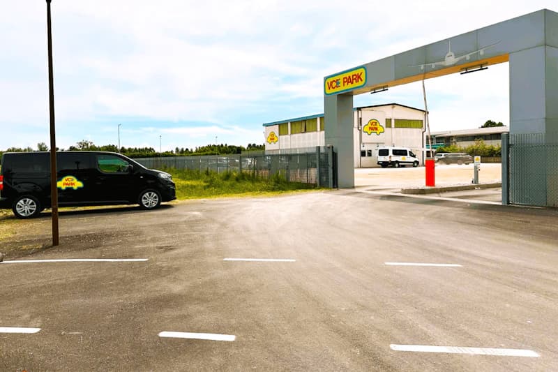 Einfahrt zu VCE PARK mit Tor und Schild, schwarzer Shuttle-Van links, leeres Asphaltareal und Gebäude im Hintergrund.