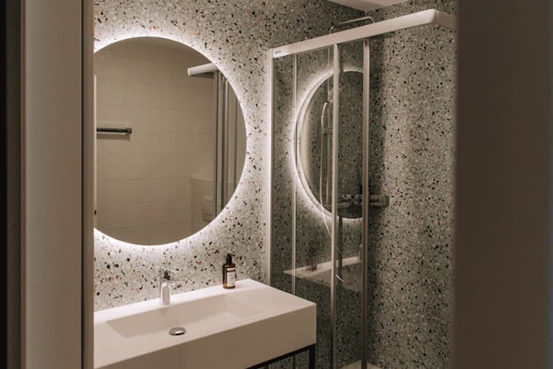 Badezimmer mit Waschbecken, rundem hinterleuchtetem Spiegel und Glasdusche vor Terrazzo-Wand.