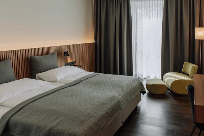 Doppelbett mit grauer Bettdecke, Holzlatten-Kopfteil, Vorhänge, Glastür und gelbe Sitzmöbel auf dunklem Holzboden.