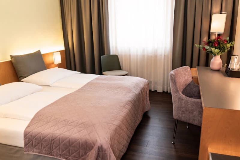 Hotelzimmer mit Doppelbett, gesteppter Bettdecke, Nachttischlampe, Schreibtisch, Sessel und Fenster mit Vorhängen.