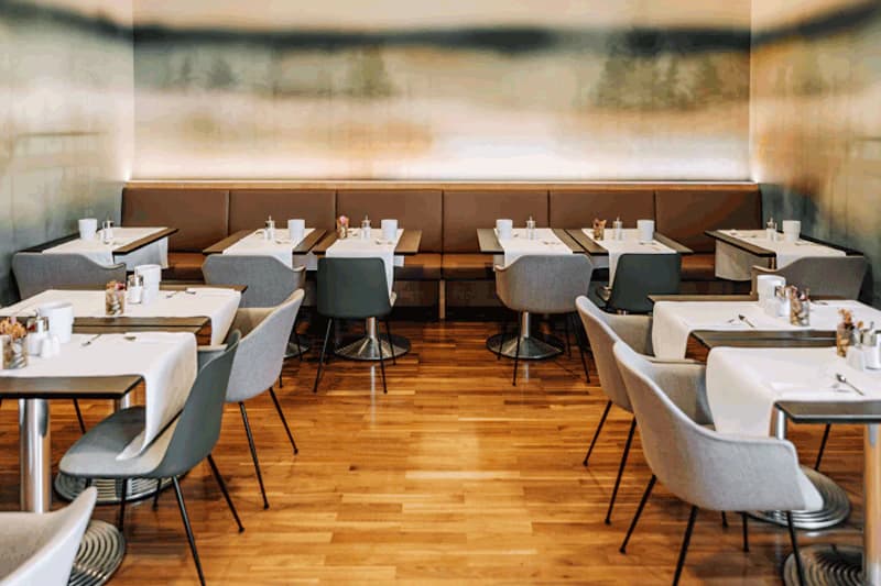Restaurantbereich mit mehreren gedeckten Tischen, grauen Polstersesseln und einer langen Sitzbank an der Wand.