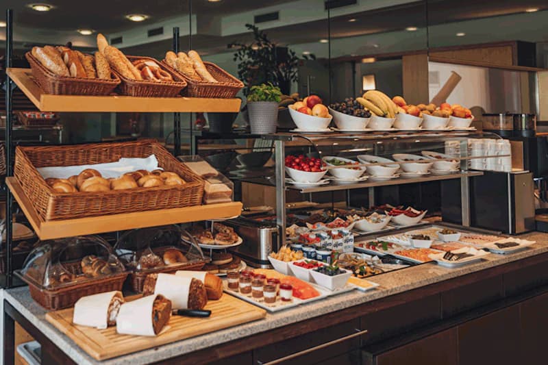 Frühstücksbuffet mit Körben voller Brötchen, Obstschalen, Aufschnitt und kleinen Joghurtgläsern.