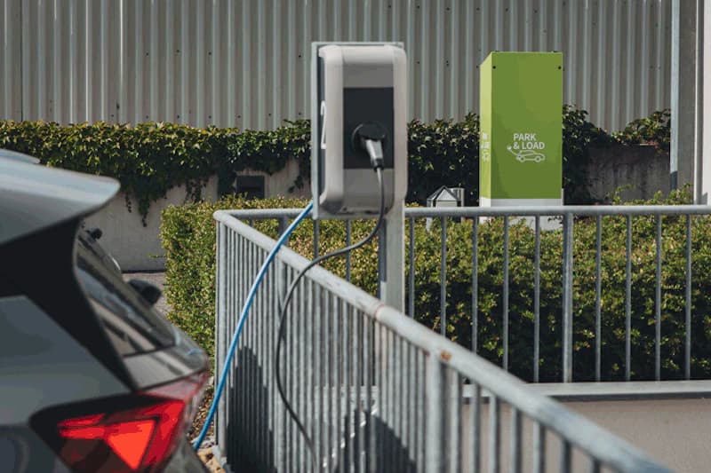 Elektroauto an Ladesäule, Kabel zum Fahrzeug, Metallgeländer und Hecke im Hintergrund.