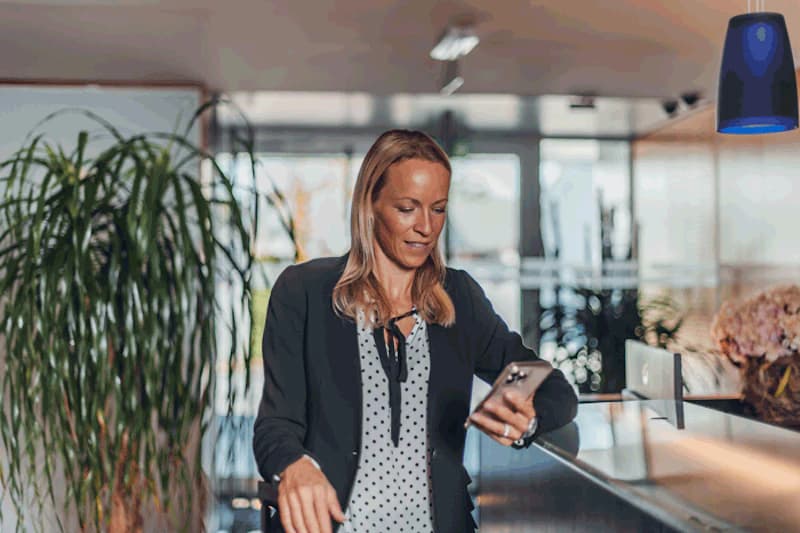 Frau an Hotelrezeption schaut auf ihr Handy, Tresen und große Zimmerpflanze im Hintergrund.