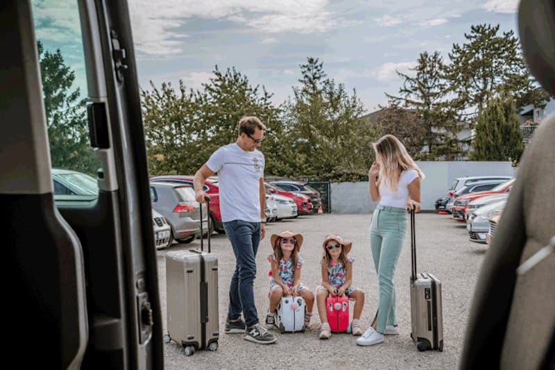 Eltern mit zwei Kindern sitzen und stehen zwischen Rollkoffern auf einem Kiesparkplatz vor geparkten Autos und Zaun.
