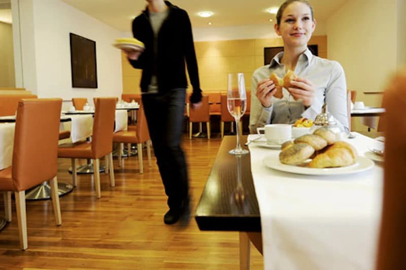 Hotelrestaurant mit Holzfußboden, Frau isst Gebäck am Tisch mit Brötchen und Sektglas, Kellner geht vorbei.