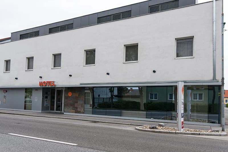 Weiße Hotelfassade mit vier kleinen Fenstern oben, Glasfront im Erdgeschoss und Eingang links mit orangefarbenem HOTEL-Schriftzug.
