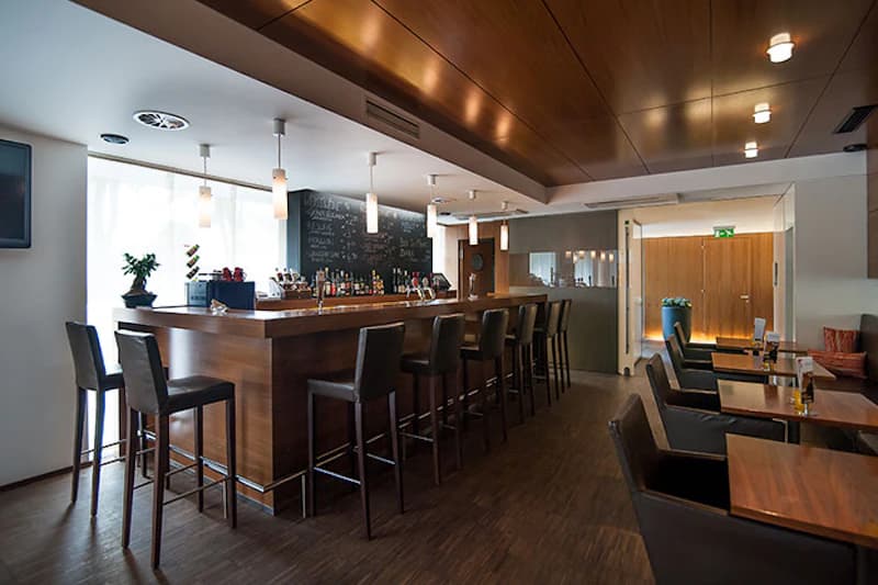 Hotelbar mit langer Holztheke, hohen Barstühlen, Tischen mit Sesseln und hängenden Leuchten.