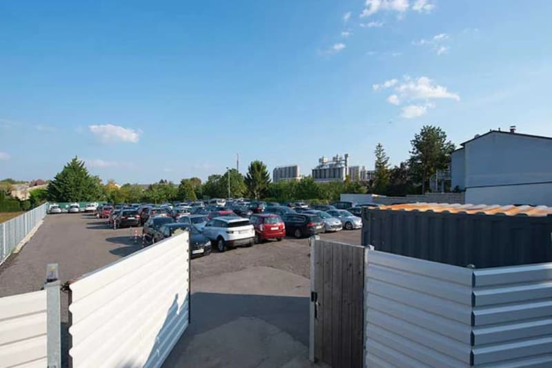 Geöffnete Einfahrt zu umzäuntem Parkplatz mit zahlreichen geparkten Autos, Wellblechcontainer und Industriegebäude im Hintergrund.