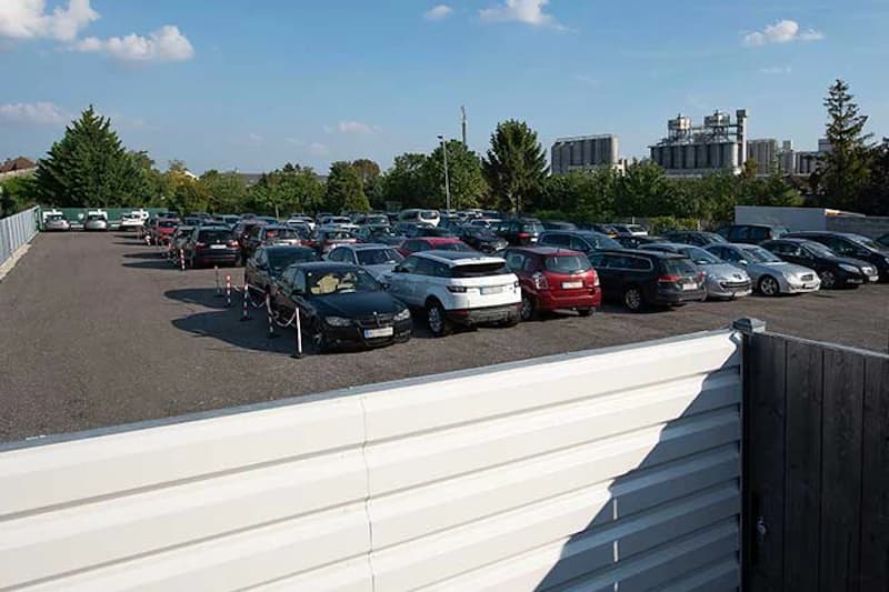 Umzäunter Parkplatz mit vielen Autos auf Schotter und weißer Metallbegrenzung im Vordergrund, Bäume und Industriegebäude im Hintergrund.