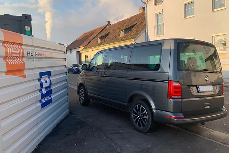 Grauer Kleinbus neben weißem Container mit HEINPARKING-Logo auf Asphaltstraße, Häuser im Hintergrund.