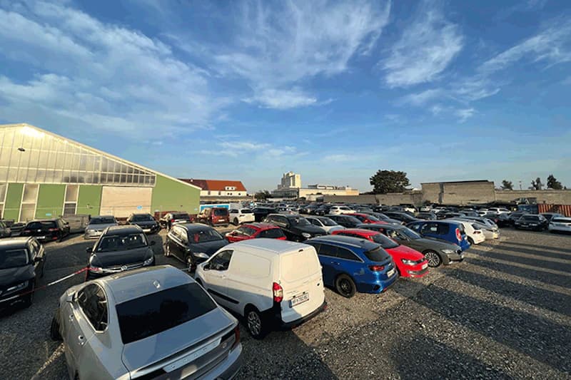 Großer Schotterparkplatz mit dicht geparkten Autos und grünem Hallengebäude unter blauem Himmel.
