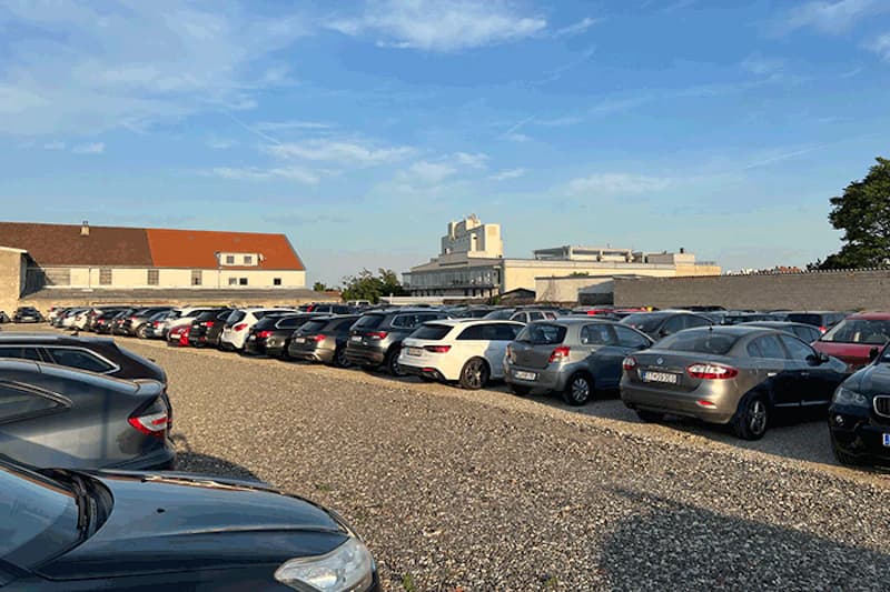 Großer Schotterparkplatz mit dicht geparkten Autos und Gebäuden im Hintergrund unter blauem Himmel.