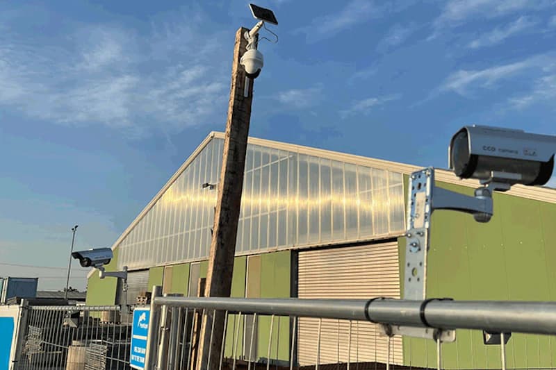 Mehrere Überwachungskameras an Zaun und Mast vor einer grünen Industriehalle bei blauem Himmel.