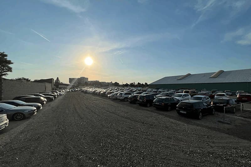 Großer Kiesparkplatz mit zwei langen Reihen geparkter Autos, Lagerhalle rechts und tief stehende Sonne am Horizont.