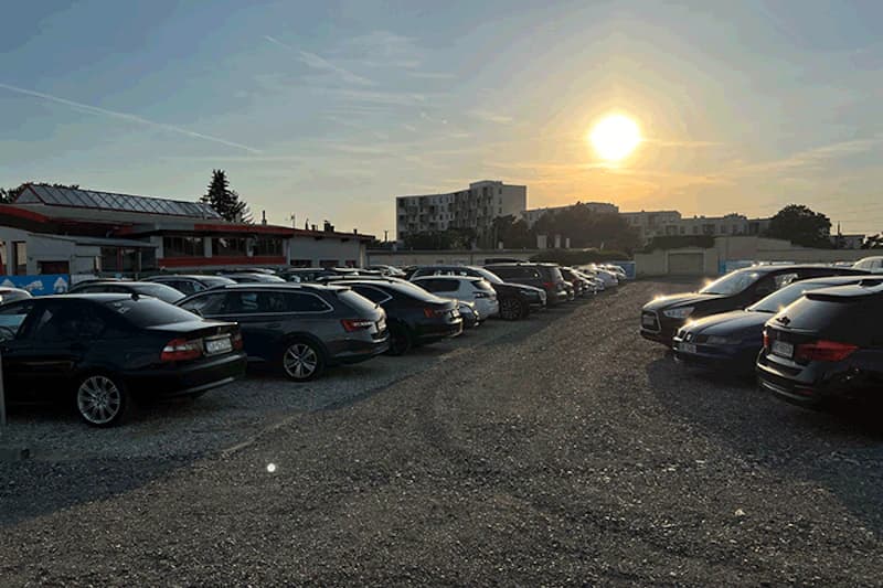 Schotterparkplatz mit Reihen geparkter Autos, niedriges Gebäude links und tief stehende Sonne am Himmel.