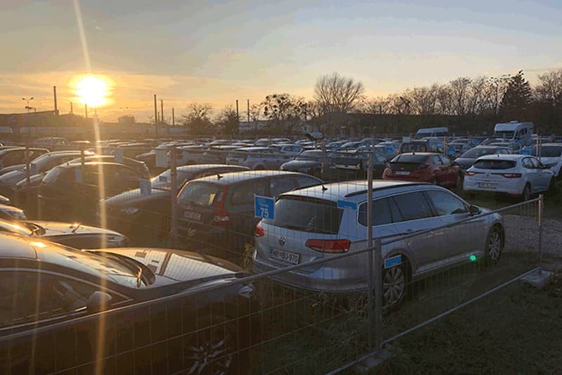 Außenparkplatz am Flughafen Wien mit umzäunten Reihen geparkter Autos bei Sonnenuntergang.