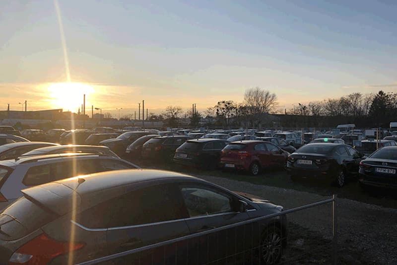 Außenparkplatz mit vielen geparkten Autos bei Sonnenuntergang, Metallzaun im Vordergrund und Industrie am Horizont.