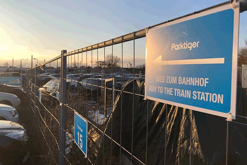Maschendrahtzaun mit Parktiger-Schild und Pfeil, dahinter Reihen geparkter Autos bei Abendlicht.