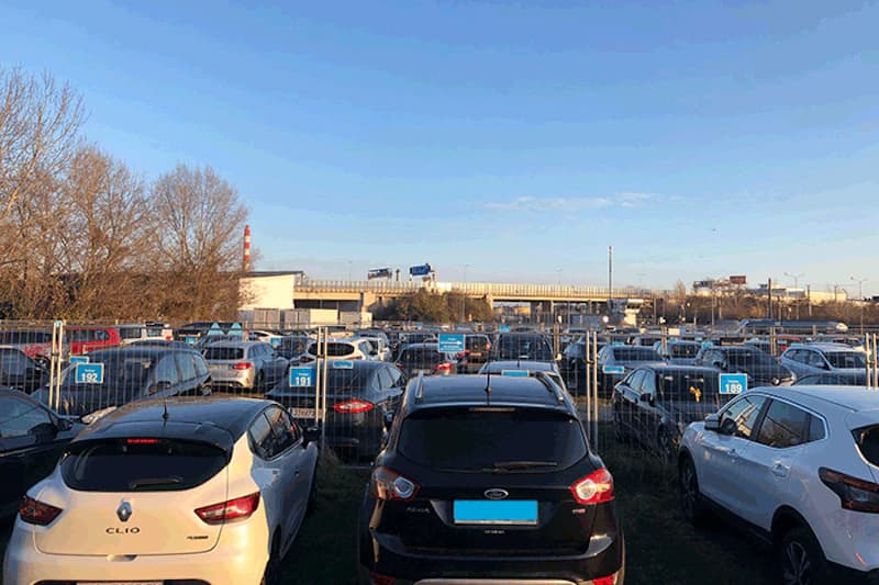 Eingezäunter Außenparkplatz mit dicht geparkten Autos und nummerierten Stellplätzen.