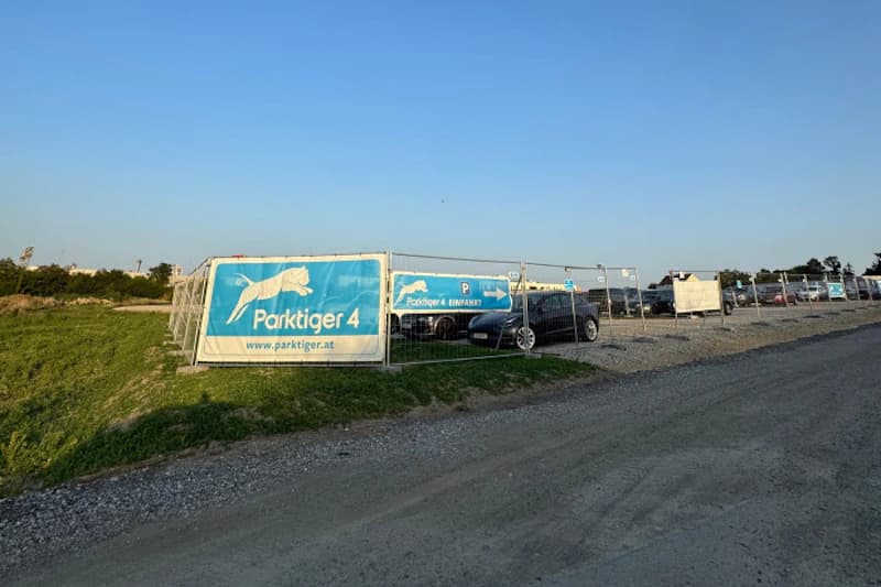 Eingezäunter Parkplatz mit Parktiger 4 Bannern, geparkten Autos und Kieszufahrt.