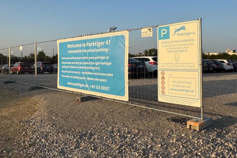 Bauzaun mit großen blauen und gelben Hinweistafeln vor Schotterparkplatz mit Reihen geparkter Autos.