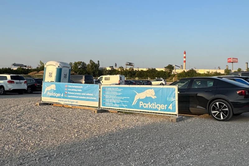 Schotterparkplatz mit blauen Parktiger 4 Bannern, mehreren geparkten Autos und einer mobilen Toilette im Abendlicht.