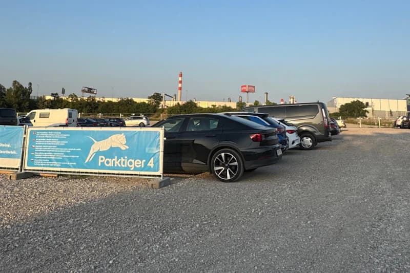 Schotterparkplatz mit geparkten Autos und blauem Parktiger 4 Banner vor Industriegebäuden bei klarem Himmel.