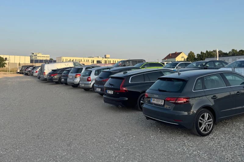 Reihung geparkter Autos auf geschottertem Parkplatz vor niedrigen Gebäuden bei klarem Himmel.