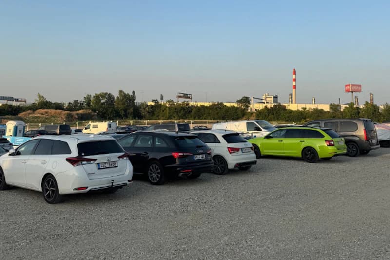 Schotterparkplatz mit mehreren geparkten Autos, einer blauen mobilen Toilette und Industrieanlagen am Horizont.
