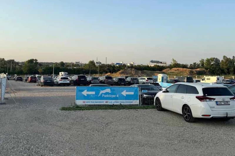 Großer Schotterparkplatz mit geparkten Autos, blaues Banner Parktiger 4 mit Richtungspfeilen und mobiler Toilette.
